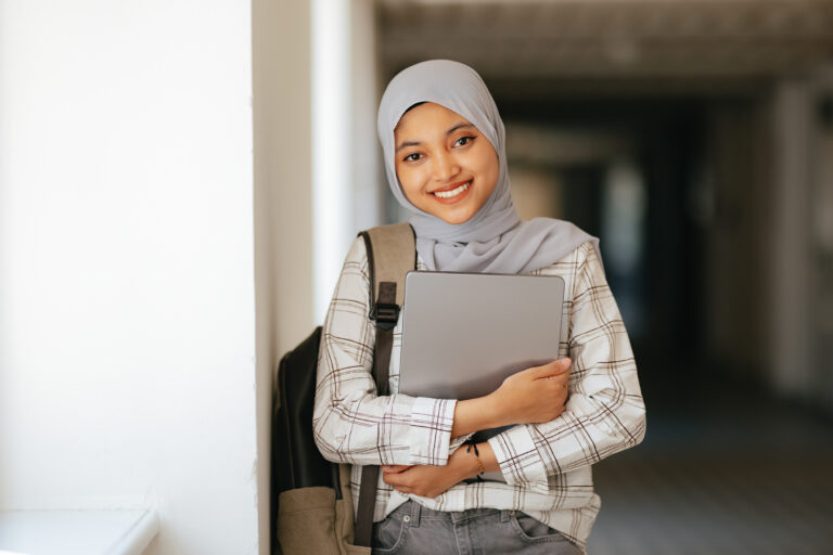 Portrait of a young female international student leaning against the wall in the school hallway. She is holding a laptop computer and backpack, smiling and looking at the camera.
